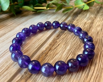 May include: A beaded bracelet composed of round, polished amethyst stones. The deep purple beads are strung together, forming a flexible band. The bracelet is displayed on a light brown wooden surface, with green foliage in the background.