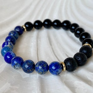 May include: A bracelet with blue lapis lazuli, black onyx, and black lava stone beads. The bracelet has gold-coloured spacers. The beads are strung on an elastic cord. The bracelet is a mix of textures and colours.