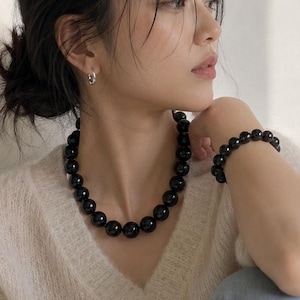 May include: A black beaded necklace and bracelet set. The necklace is a choker style with large, round beads. The bracelet has a similar design. The jewelry is worn with a cream-coloured sweater and blue jeans.