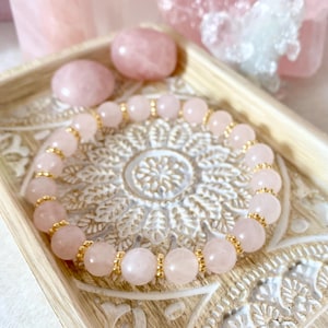 May include: A rose quartz beaded bracelet with gold-coloured accents rests on a decorative wooden tray. Two rose quartz stones and other pink crystals are in the background. The bracelet features round, light pink beads separated by gold-coloured spacers.