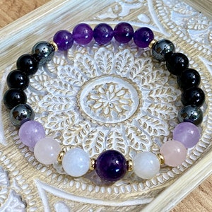 May include: A purple, black, and white beaded bracelet with gold spacers. The bracelet is made with amethyst, black onyx, hematite, rose quartz, and moonstone beads.