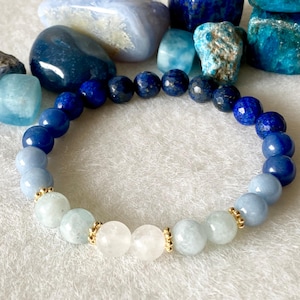 May include: A beaded bracelet featuring a gradient of blue and white gemstones. The bracelet includes light blue, medium blue, and dark blue beads, with white and gold accents. The bracelet is displayed with various blue gemstones in the background.