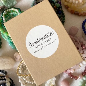 May include: A rectangular brown cardboard gift box with a circular white sticker featuring the black text Apartment020 GEM & SILVER Jewelry for your soul, surrounded by various colorful gemstone bead bracelets and loose polished stones.