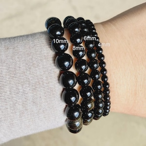 May include: Four black beaded bracelets featuring smooth, spherical stones in varying sizes. The bracelets are labeled with text indicating bead diameters of 10mm, 8mm, 6mm, and 4mm, showcasing the different stone sizes available for these accessories.