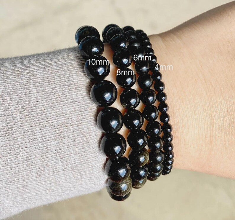 May include: Four black beaded bracelets featuring smooth, spherical stones in varying sizes. The bracelets are labeled with text indicating bead diameters of 10mm, 8mm, 6mm, and 4mm, showcasing the different stone sizes available for these accessories.