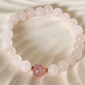 Pu&ograve; includere: Un braccialetto di pietre bianche con un unico ciondolo rosa al centro. Il braccialetto &egrave; realizzato con dettagli in oro rosa.