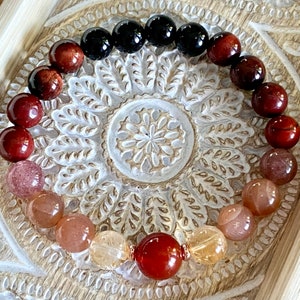 May include: A multi-colored gemstone bracelet with red, orange, and black beads. The bracelet is made of natural stones and is arranged in a gradient pattern.