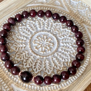 May include: A red gemstone bracelet with a rose gold clasp. The bracelet is made of round beads and is lying on a decorative wooden surface.