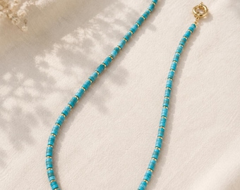 May include: A turquoise beaded necklace with gold-coloured accents. The necklace features small, rectangular turquoise beads separated by small gold-coloured cylindrical beads. The necklace is displayed on a white surface.