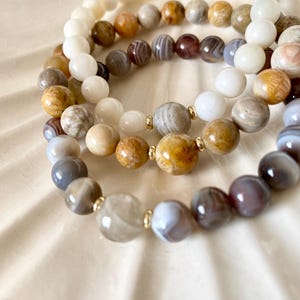 May include: Four beaded bracelets in shades of white, brown, and grey. The bracelets are made of round, polished beads, with gold-coloured accents. The bracelets are arranged in a curved pattern, showcasing the variety of colours and textures.