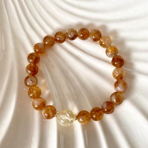 Peut inclure: Un bracelet élastique composé de perles rondes et translucides en quartz rutile doré. Les perles présentent des inclusions internes en forme d'aiguilles dans des tons ambre et miel. Une perle de quartz rutile jaune pâle est centrée.