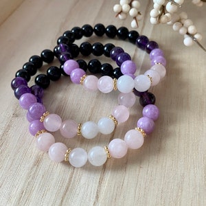 May include: Three elastic bracelets featuring smooth, round beads. One bracelet consists of black and deep purple beads. The other two bracelets feature a gradient of light purple, soft pink, and white beads, accented with small gold-toned metal spacers.