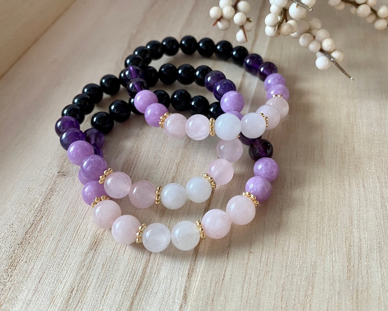 May include: Three elastic bracelets featuring smooth, round beads. One bracelet consists of black and deep purple beads. The other two bracelets feature a gradient of light purple, soft pink, and white beads, accented with small gold-toned metal spacers.