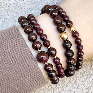 May include: Two stacked bracelets featuring deep red, polished spherical beads. The bracelets are accented with small, shiny gold-toned metal spacer beads and one larger, smooth gold-toned metal bead positioned centrally on the outer strand.