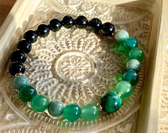 Puede incluir: Un brazalete de piedras preciosas negras y verdes con un acento dorado. El brazalete está hecho de cuentas redondas y presenta una variedad de tonos verdes.