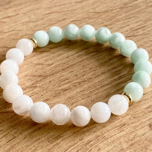 May include: A white and light green gemstone bracelet with gold accents. The bracelet is made of round beads and is approximately 18 cm in length.