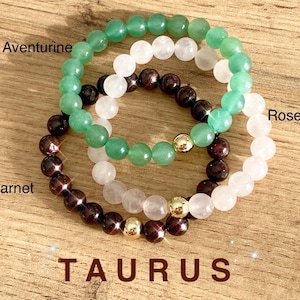 May include: Three beaded bracelets in green aventurine, rose quartz, and garnet. The bracelets are stacked on top of each other and have gold beads separating the stones. The text "TAURUS" is written below the bracelets.