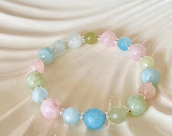 Könnte beinhalten: Ein Armband mit Perlen aus pastellfarbenen Edelsteinen, darunter Rosa, Blau und Grün. Die Perlen sind durch silberfarbene Abstandshalter getrennt. Das Armband liegt auf einer weißen, strukturierten Oberfläche.