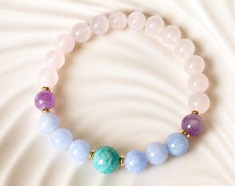 Könnte beinhalten: Ein Armband mit Edelsteinperlen. Das Armband besteht aus hellrosa Rosenquarz, lila Amethyst, hellblauen Edelsteinen und einem grünen Edelstein. Goldfarbene Abstandshalter trennen die verschiedenen Edelsteinarten. Das Armband wird auf einer weißen Oberfläche präsentiert.