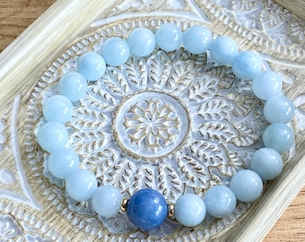 Könnte beinhalten: Ein hellblaues Edelstein-Armband mit einem größeren, runden, hellblauen Edelstein-Perlen in der Mitte. Das Armband liegt auf einer weißen, dekorativen Oberfläche.