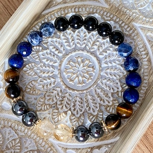 May include: A beaded bracelet featuring a circular arrangement of faceted and smooth gemstone beads in shades of black, deep blue, brown, and a pair of clear yellow beads at the bottom, accented with small gold-toned metal spacers throughout.