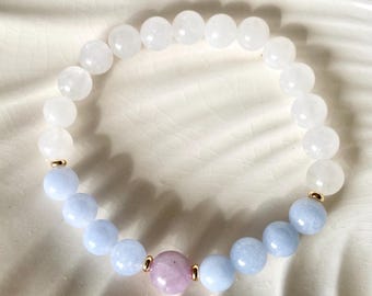 Könnte beinhalten: Ein Armband mit weißen, hellblauen und einer einzelnen rosa Edelsteinperle. Das Armband hat goldfarbene Akzente. Die Perlen sind auf einer elastischen Schnur aufgereiht. Das Armband wird auf einer weißen, strukturierten Oberfläche präsentiert.