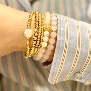 May include: A selection of gold beaded and rose quartz bracelets. One bracelet has a large white pearl. The bracelets are stacked on a wrist, against a light blue striped shirt.