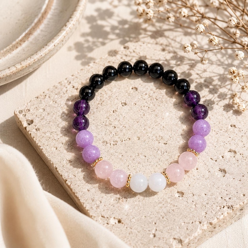 May include: A beaded bracelet featuring a gradient of smooth, round gemstone beads. The color sequence transitions from black to deep purple, light purple, soft pink, and finally white, accented by small gold-toned metal spacer beads.