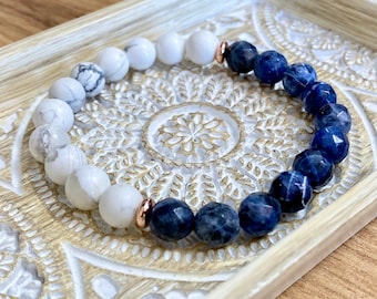 Puede incluir: Un brazalete de piedras preciosas blancas y azules con detalles dorados. El brazalete está hecho de cuentas blancas de howlite y cuentas azules de sodalita.