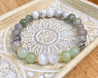 May include: A multi-colored beaded bracelet with white, gray, green, and black beads. The bracelet is made with natural stones and has gold accents.