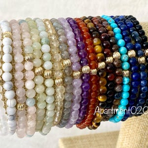 May include: A stack of 12 colorful beaded bracelets with gold accents. The bracelets are made of various stones, including white, pink, green, purple, red, brown, blue, and black.