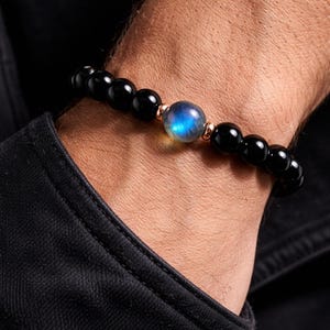 May include: Black beaded bracelet featuring a central blue-green iridescent stone and rose gold details. The bracelet is composed of round, polished black beads. The central stone is a round, iridescent blue-green colour. The bracelet is worn on a wrist.