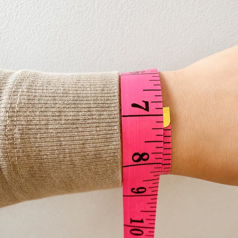 May include: A beige ribbed long-sleeve garment cuff positioned next to a bright pink measuring tape. The measuring tape shows a circumference measurement of approximately 7 inches around the wrist area, clearly displaying the numerical markings.