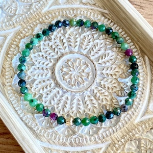 May include: A beaded bracelet with green, purple, and dark blue gemstones. The bracelet has a gold-coloured clasp. The bracelet is displayed on a carved wooden tray with a floral design. The gemstones are faceted.