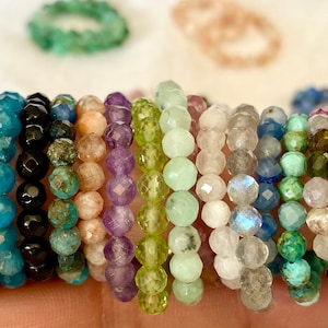 May include: A stack of 15 gemstone bracelets, each made with faceted beads. The bracelets are in various colours, including green, blue, purple, and brown.