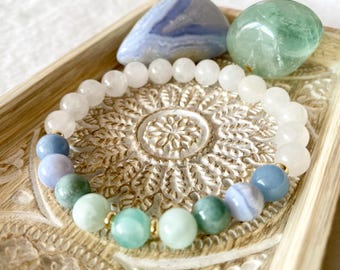 Puede incluir: Un brazalete hecho con cuentas de piedras preciosas blancas, azules y verdes. El brazalete está en una bandeja de madera con un diseño decorativo. También hay dos piedras preciosas más grandes en la bandeja, una azul y una verde.