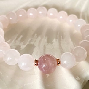Pu&ograve; includere: Un braccialetto di perline rosa e bianche con una perla rosa pi&ugrave; grande al centro. Il braccialetto &egrave; fatto di pietre naturali e ha un accento oro rosa.