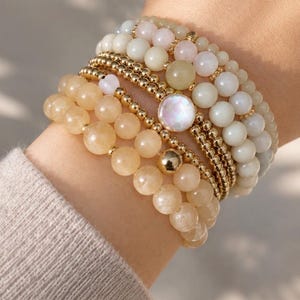 May include: A stack of beaded bracelets featuring round polished stones in shades of peach, cream, and pale pink, interspersed with small metallic gold beads and a single larger central gold bead, plus one bracelet with a round iridescent charm.