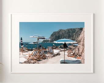 May include: A framed photograph of a beach scene with blue and white striped beach chairs and umbrellas. The beach is sandy and there are rocks in the background.