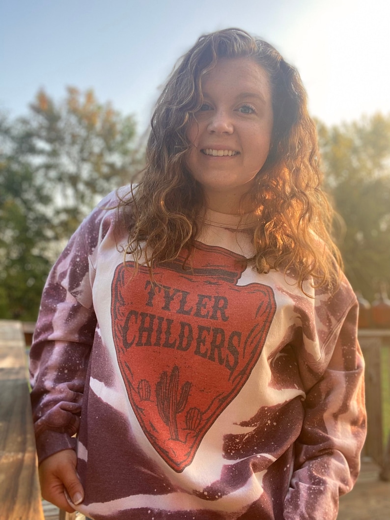 May include: A tie-dye sweatshirt in shades of purple and white with a red and white graphic of a vintage arrow with the text "Tyler Childers" inside.