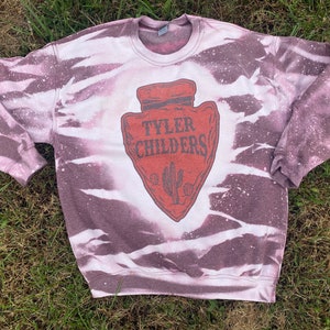 May include: A white and purple tie-dye sweatshirt with a red arrowhead graphic featuring the text "Tyler Childers".