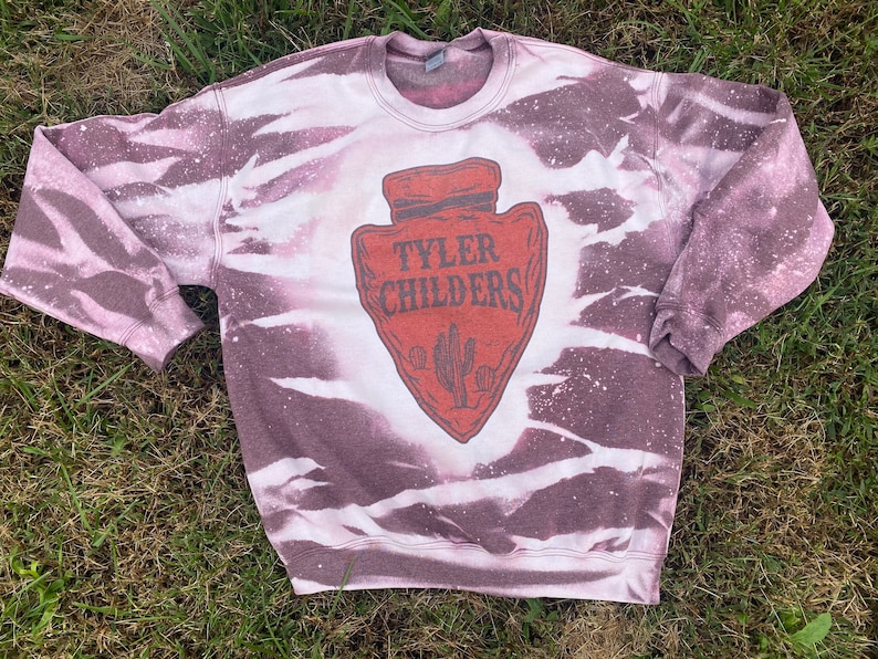 May include: A white and purple tie-dye sweatshirt with a red arrowhead graphic featuring the text "Tyler Childers".