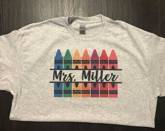 May include: A light gray t-shirt featuring a colorful crayon design. The design includes crayons in various colors, with the name "Mrs. Miller" printed in black script below the crayons. The shirt is a Gildan brand.