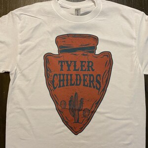 May include: White t-shirt with a red arrowhead design featuring the text "Tyler Childers" in blue. The arrowhead has a distressed look and includes a cactus graphic.