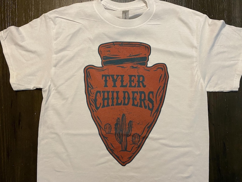 May include: White t-shirt with a red arrowhead design featuring the text "Tyler Childers" in blue. The arrowhead has a distressed look and includes a cactus graphic.