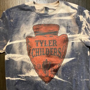 May include: A blue and white tie-dye t-shirt with a red arrowhead design. The arrowhead has the text "Tyler Childers" in white lettering.
