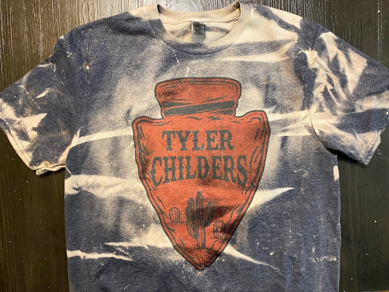 May include: A blue and white tie-dye t-shirt with a red arrowhead design. The arrowhead has the text "Tyler Childers" in white lettering.