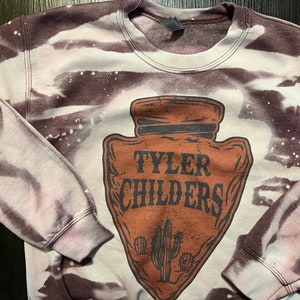 May include: A brown and white tie-dye sweatshirt with a brown arrowhead design featuring the text "Tyler Childers" and a cactus graphic.