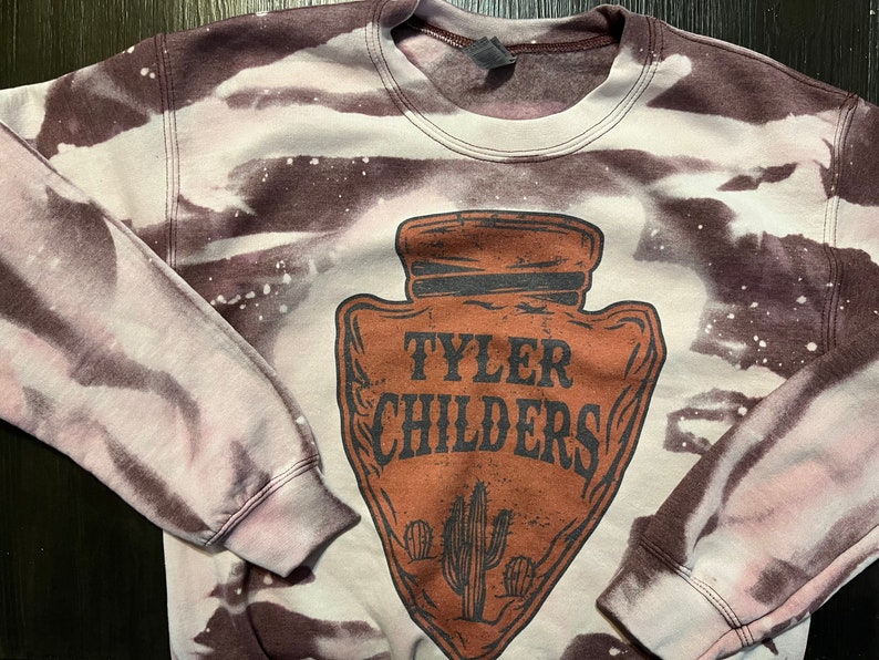 May include: A brown and white tie-dye sweatshirt with a brown arrowhead design featuring the text "Tyler Childers" and a cactus graphic.