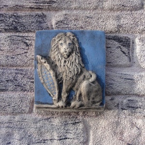 May include: A square, blue and grey relief sculpture of a lion holding a shield. The lion has a detailed mane and blue eyes. The shield has a blue and white pattern. The sculpture is mounted on a textured brick wall.